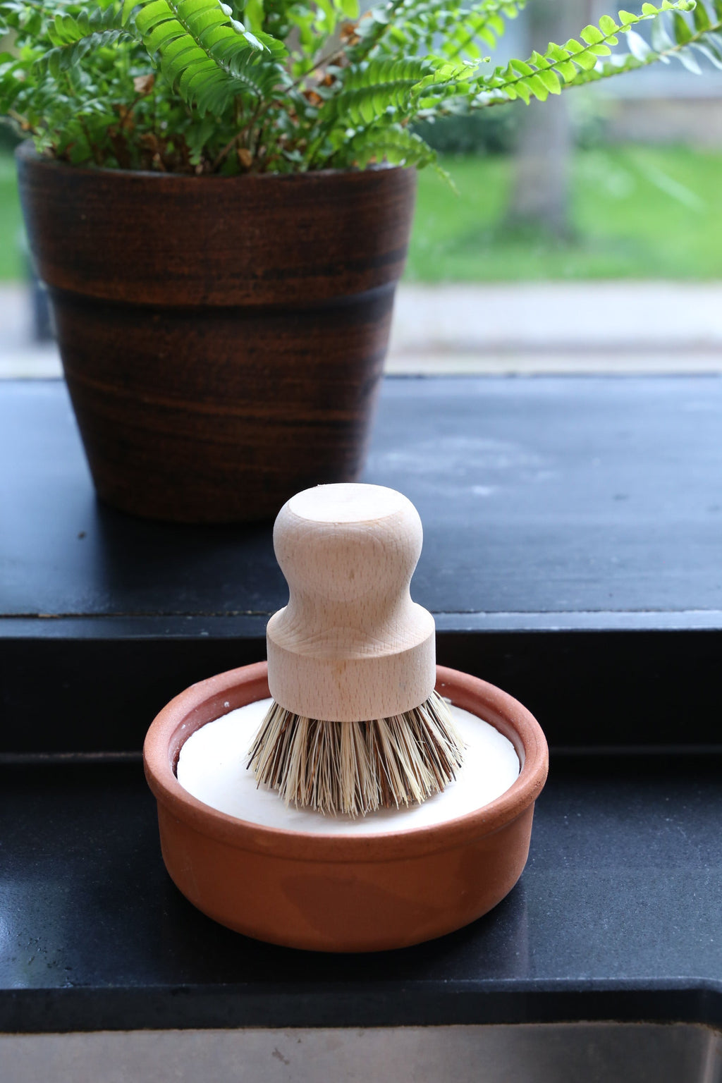Cleaning brush with wooden handle and terracotta base and solid dishwashing soap on a windowsill with a plant in the background 